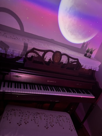 An elegant upright piano with a lace cover is placed in a room with a surreal projection of a large moon on the wall. The colors of the projection cast a soft pink and purple glow over the scene, and a small vase with plants is situated on top of the piano.