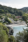 A coastal village nestled among lush, green hills with a pebbled beach and clear blue waters. Several houses and structures with terracotta roofs are visible, surrounded by dense vegetation including tall cypress and pine trees. The beach features sun loungers and umbrellas suggesting a popular spot for relaxation.