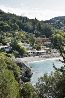 A coastal village nestled among lush, green hills with a pebbled beach and clear blue waters. Several houses and structures with terracotta roofs are visible, surrounded by dense vegetation including tall cypress and pine trees. The beach features sun loungers and umbrellas suggesting a popular spot for relaxation.