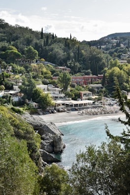 A coastal village nestled among lush, green hills with a pebbled beach and clear blue waters. Several houses and structures with terracotta roofs are visible, surrounded by dense vegetation including tall cypress and pine trees. The beach features sun loungers and umbrellas suggesting a popular spot for relaxation.
