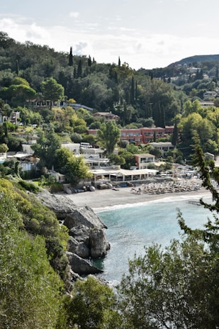A coastal village nestled among lush, green hills with a pebbled beach and clear blue waters. Several houses and structures with terracotta roofs are visible, surrounded by dense vegetation including tall cypress and pine trees. The beach features sun loungers and umbrellas suggesting a popular spot for relaxation.