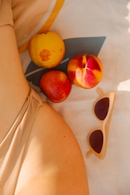 Three ripe peaches are placed on a patterned cloth next to a pair of stylish sunglasses. The lower back of a person wearing a textured beige swimsuit is partially visible, creating a relaxed and summery vibe.