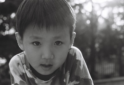 A young child with a serious expression is gazing directly at the camera. The image is in black and white, creating a nostalgic and timeless feel. The background is blurred with soft bokeh effects, suggesting a natural outdoor setting.