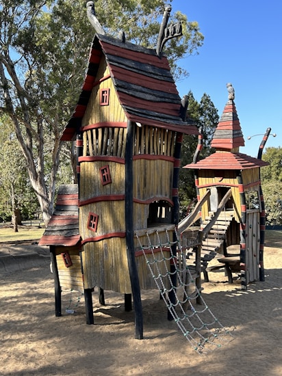 A whimsical and rustic playground structure resembling a fantasy house, constructed with wooden materials and featuring slanted roofs, ropes, and slides. The structure includes bright red accents and black detailing, situated on a sandy ground with trees in the background, creating a fairy-tale like atmosphere.