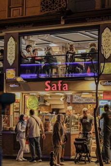 A two-level cafe or restaurant with the name 'Safa'. The upper floor has people sitting at tables, engaged in conversation. The ground level features a display counter with food and customers standing in front. It is evening and the place is warmly lit, creating a cozy atmosphere.