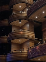 Wooden balconies with curved designs and dark railings, illuminated by soft lighting with spotlights. The architecture features layers of elegantly crafted wood panels.