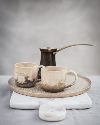 Two rustic ceramic mugs with a speckled, earthy texture are placed on a round tray. Behind the mugs, an aged metal coffee pot with a long handle stands out. The setup rests on a white marble board against a blurred grey background, creating a serene and minimalistic composition.