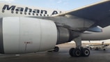 Close-up view of an airplane's engine and landing gear on the tarmac. The logo of an airline is visible on the body of the aircraft. In the background, there are service vehicles.