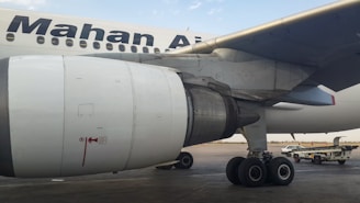 Close-up view of an airplane's engine and landing gear on the tarmac. The logo of an airline is visible on the body of the aircraft. In the background, there are service vehicles.