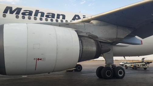 Close-up view of an airplane's engine and landing gear on the tarmac. The logo of an airline is visible on the body of the aircraft. In the background, there are service vehicles.