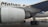 Close-up view of an airplane's engine and landing gear on the tarmac. The logo of an airline is visible on the body of the aircraft. In the background, there are service vehicles.