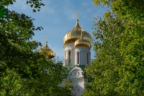 Golden domes of an Orthodox church rise above lush green trees, creating a blend of natural and architectural beauty under a clear blue sky.