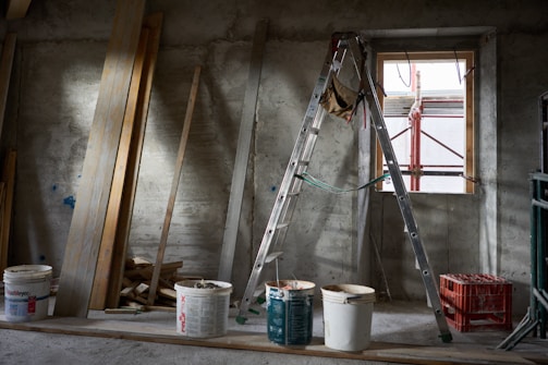 A construction site room features a metal ladder positioned near a window with visible scaffolding. Planks of wood are leaned against a concrete wall, and several paint buckets are scattered on the floor, with one bucket holding a trowel. The space is dimly lit, casting shadows around.