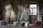 A construction site room features a metal ladder positioned near a window with visible scaffolding. Planks of wood are leaned against a concrete wall, and several paint buckets are scattered on the floor, with one bucket holding a trowel. The space is dimly lit, casting shadows around.