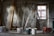 A construction site room features a metal ladder positioned near a window with visible scaffolding. Planks of wood are leaned against a concrete wall, and several paint buckets are scattered on the floor, with one bucket holding a trowel. The space is dimly lit, casting shadows around.