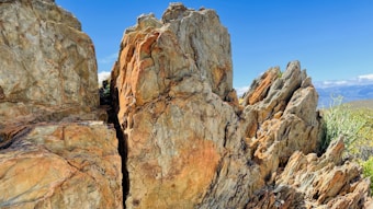 Large rugged rocks with sharp edges and varying earth tones are prominently featured in an outdoor setting. The background consists of clear blue skies and distant mountains, suggesting a natural, mountainous environment.
