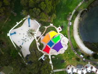 Aerial view of a colorful playground with multiple vibrant shade structures in a park setting. The structures include orange, yellow, green, blue, and purple triangular canopies. The playground features various play equipment and is surrounded by pathways and greenery. Nearby, a body of water and additional parkland can be seen.