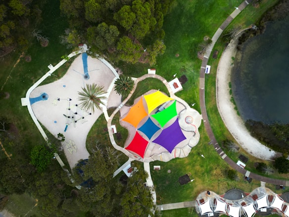 Aerial view of a colorful playground with multiple vibrant shade structures in a park setting. The structures include orange, yellow, green, blue, and purple triangular canopies. The playground features various play equipment and is surrounded by pathways and greenery. Nearby, a body of water and additional parkland can be seen.