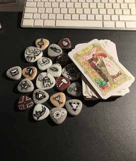 A collection of stones with various symbols painted on them, accompanied by a deck of illustrated tarot-style cards. The symbols include abstract drawings like eyes, hearts, and arrows, painted in black on white and beige stones. The cards feature colorful artwork with one visible card depicting a couple embracing with the word 'Joie' at the top. Everything is placed on a dark surface, with a part of a computer keyboard visible in the background.