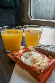 Two plastic cups filled with orange juice are placed on a tray table. Alongside them, there are packets of cookies, with one labeled as salted caramel flavored. The setting appears to be on a train, with a window and seat visible in the background.