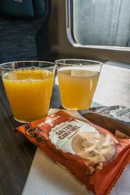 Two plastic cups filled with orange juice are placed on a tray table. Alongside them, there are packets of cookies, with one labeled as salted caramel flavored. The setting appears to be on a train, with a window and seat visible in the background.