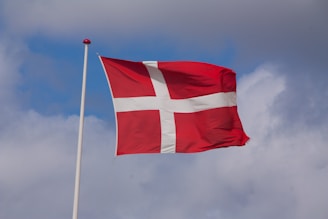 A red flag with a white Nordic cross is waving against a partly cloudy sky.