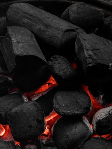 A close-up of hot charcoal and wood pieces glowing with intense red-orange embers in between dark black chunks. The surface texture is rough, a mix of irregular cylindrical and cuboidal shapes, with visible cracks and crevices in the material.