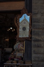 A decorated mirror outside a shop reflects the facade of a historic building with intricate architectural details, including a rose window and triangular pediment. The shop has various ornate items on display, such as trays and decorative objects.
