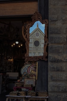 A decorated mirror outside a shop reflects the facade of a historic building with intricate architectural details, including a rose window and triangular pediment. The shop has various ornate items on display, such as trays and decorative objects.