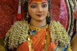A colorful statue adorned with intricate jewelry and floral garlands. The face is painted with elaborate makeup, emphasizing the eyes and forehead, and surrounded by detailed accessories. The figure wears a vibrant sari, combining orange and blue hues, and stands against a shimmering background.