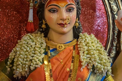 A colorful statue adorned with intricate jewelry and floral garlands. The face is painted with elaborate makeup, emphasizing the eyes and forehead, and surrounded by detailed accessories. The figure wears a vibrant sari, combining orange and blue hues, and stands against a shimmering background.