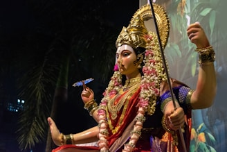 A beautifully crafted statue of a deity adorned with elaborate jewelry, vibrant clothing, and a golden headdress. The figure is holding a long staff and a floral garland, and is surrounded by flowers. The background features lush greenery, contributing to a serene and divine ambiance.