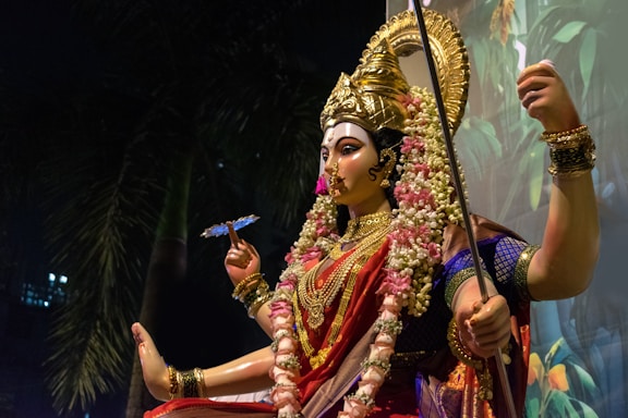 A beautifully crafted statue of a deity adorned with elaborate jewelry, vibrant clothing, and a golden headdress. The figure is holding a long staff and a floral garland, and is surrounded by flowers. The background features lush greenery, contributing to a serene and divine ambiance.
