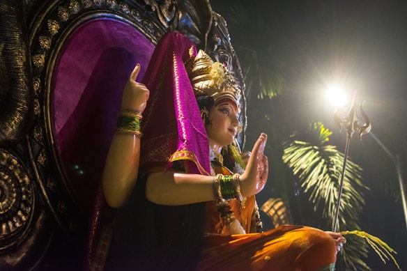 A detailed sculpture depicts a multi-armed deity adorned with vibrant garments and jewelry, positioned against a decorative backdrop. A trident is held in one of the deity’s hands, and there is an ambient glow from a nearby light source, casting dramatic shadows on the scene.