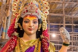 A colorful and intricately designed statue of a deity adorned with a vibrant saree and ornate jewelry. The statue features an elaborate headdress decorated with jewels and floral garlands. The backdrop consists of bamboo scaffolding under bright lights, suggesting a festival or celebration setting.