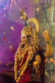 A colorful and ornate statue of a deity adorned with intricate jewelry and flowers. The figure has a golden headdress and holds a trident and another object in its hands. Brightly colored garments and a garland of flowers add to the festive appearance against a vibrant backdrop.