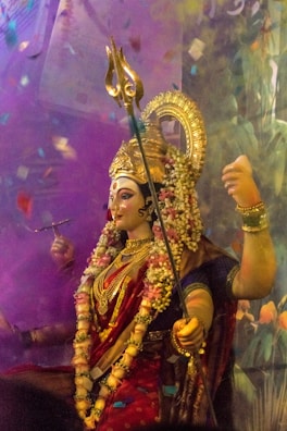 A colorful and ornate statue of a deity adorned with intricate jewelry and flowers. The figure has a golden headdress and holds a trident and another object in its hands. Brightly colored garments and a garland of flowers add to the festive appearance against a vibrant backdrop.