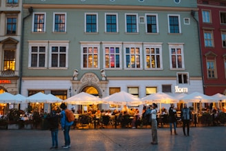 An outdoor dining area bustling with people under large, white umbrellas in front of a multi-story, historic-looking building. Warm lighting emanates from the restaurant, and several people stand and walk around the cobblestone plaza. The building features large windows and decorative elements.