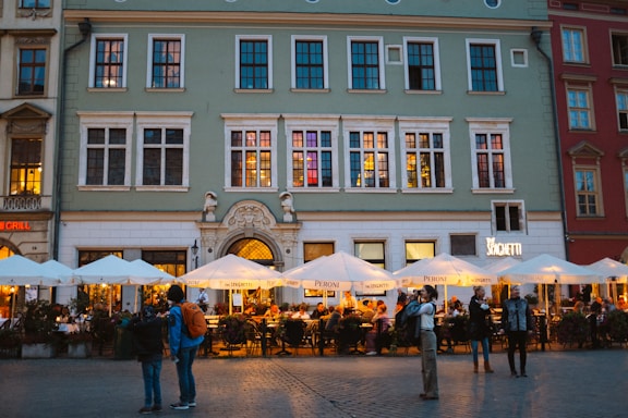 An outdoor dining area bustling with people under large, white umbrellas in front of a multi-story, historic-looking building. Warm lighting emanates from the restaurant, and several people stand and walk around the cobblestone plaza. The building features large windows and decorative elements.