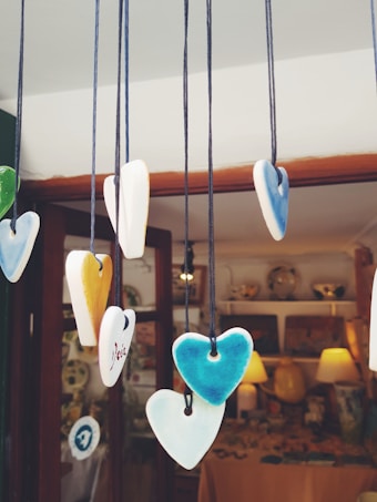 Colorful ceramic heart-shaped ornaments hang from strings inside a shop. The hearts are painted in various shades including green, blue, white, and yellow. The background shows a warm, cozy interior with soft lighting and decorative items displayed.