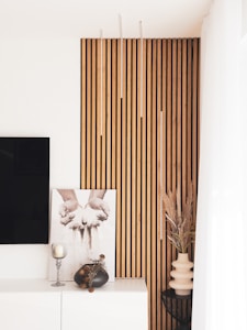 Modern interior design featuring a vertical wooden slat wall accent, a framed black-and-white photograph of hands releasing sand, and minimalistic decor including a candle holder and vases with dried plants. Light and airy with a contemporary aesthetic.