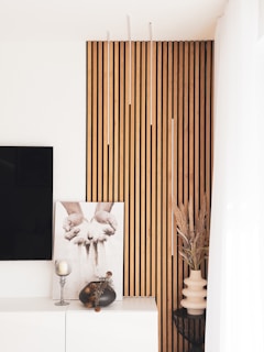 Modern interior design featuring a vertical wooden slat wall accent, a framed black-and-white photograph of hands releasing sand, and minimalistic decor including a candle holder and vases with dried plants. Light and airy with a contemporary aesthetic.