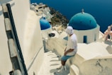A person wearing a white hat and striped shirt sits on a narrow stairway overlooking a picturesque coastal village. The buildings are predominantly white with blue-domed roofs, typical of Cycladic architecture. The deep blue sea is visible in the background, merging seamlessly with the clear sky.