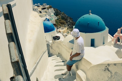 A person wearing a white hat and striped shirt sits on a narrow stairway overlooking a picturesque coastal village. The buildings are predominantly white with blue-domed roofs, typical of Cycladic architecture. The deep blue sea is visible in the background, merging seamlessly with the clear sky.