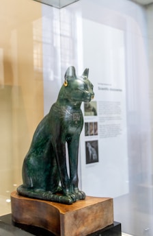 A bronze statue of a sitting cat, adorned with intricate engravings and wearing gold earrings, is placed on a wooden base. The background shows blurred text and images on a wall, indicating a museum exhibit setting.