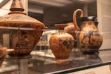A collection of ancient pottery items displayed in a museum setting, featuring intricate designs and patterns. The pottery includes a large bowl with a lid, a small vase, and a jug, all adorned with animal motifs and floral patterns.
