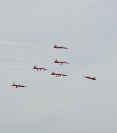 A formation of six red jets is flying in a coordinated pattern against a gray sky. The jets are leaving white smoke trails behind them as they maneuver in the air.