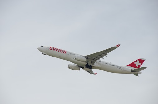 A commercial passenger airplane in mid-flight with the branding 'SWISS' prominently displayed on its body. The tail features the distinctive red and white cross logo. The aircraft is flying against a backdrop of a clear, pale sky.