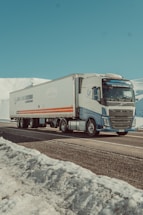 A large truck is driving along a snowy mountain road. The truck is mostly white with blue and orange detailing and features a long trailer. Snow-covered mountains and a clear blue sky dominate the background, creating a serene and calm landscape.
