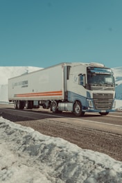 A large truck is driving along a snowy mountain road. The truck is mostly white with blue and orange detailing and features a long trailer. Snow-covered mountains and a clear blue sky dominate the background, creating a serene and calm landscape.